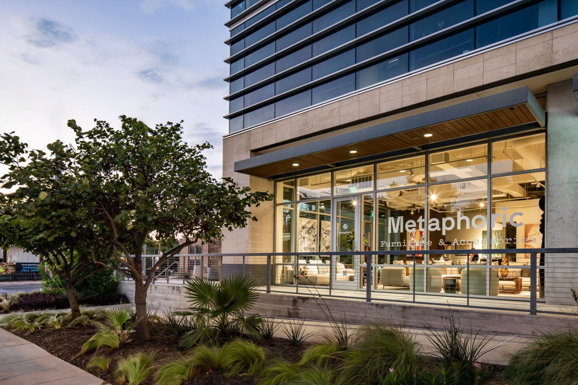 Metaphoric Furniture & Accessories storefront with floor-to-ceiling windows at ground level of a modern glass high-rise building at dusk